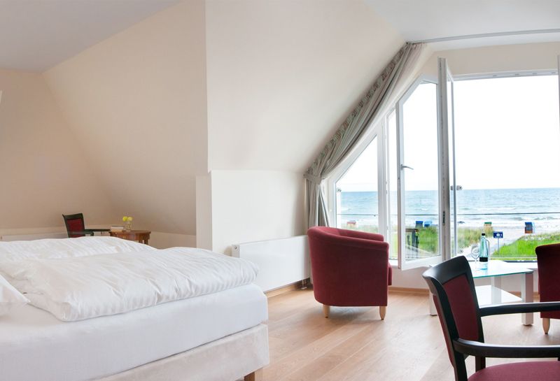Blick vom Bett aus auf die Ostsee. Die großen Fenster stehen auf und vor ihnen stehen zwei rote Sessel