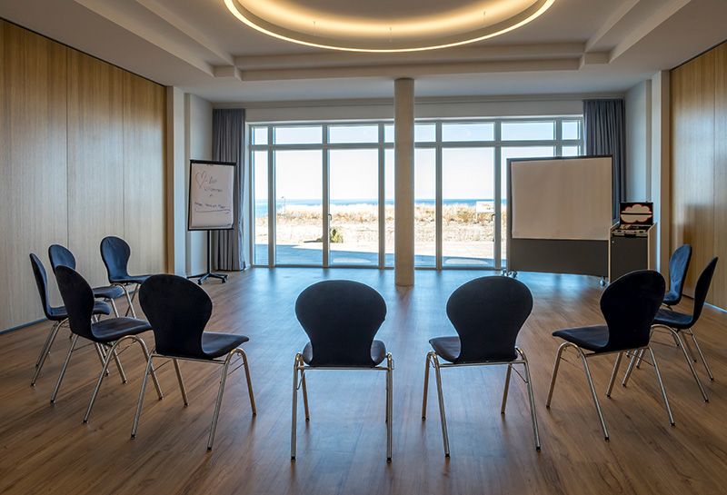 Tagen am Meer, Tagungsraum, Stuhlkreis, blaue Stühle, vor den Stühlen Flipchart, Pinnwand auf Füßen, Rollcontainer mit Tagungsutensilien, Fensterfront mit Blick auf die Sonnenterrasse und Ostsee
