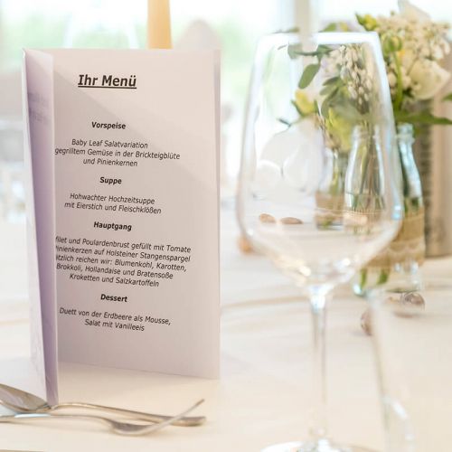 Feierlichkeiten Haus am Meer, Nahaufnahme feierlich geschmückter Tisch, weiße Tischdecke, Weinglas, Besteck, Vase mit Blumenschmuck, aufgeklappte Menükarte