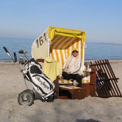 Aktivitäten Hohwachter Bucht, am Strand, geöffneter gelb-weiß gestreifter Strandkorb, Mann sitzt im Strandkorb, links neben dem Strandkorb Golfbag mit Schlägern, im Hintergrund Ostsee
