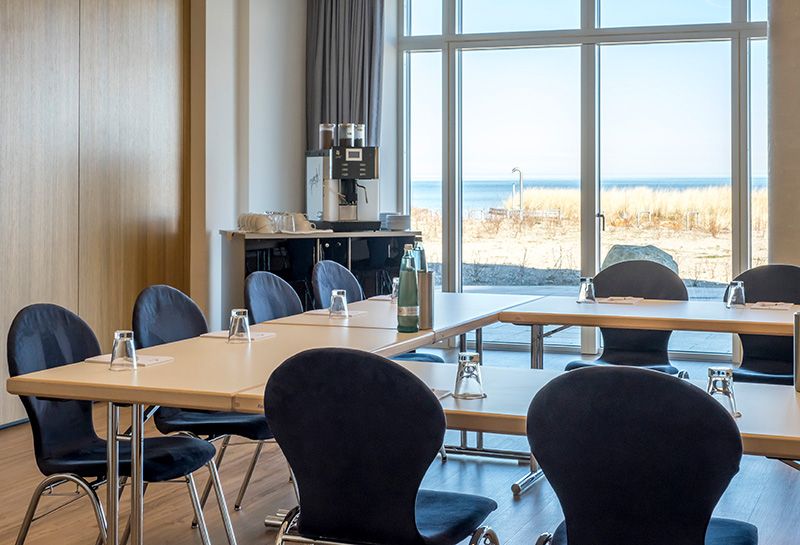 Tagen Hotel Haus am Meer, Tagungsraum, Bestuhlung U-Form, Tische gedeckt mit Wassergläsern, Wasserflaschen, Blöcken, in der linken Ecke Rollwagen mit Kaffeevollautomat und Tassen, Blick auf die Ostsee