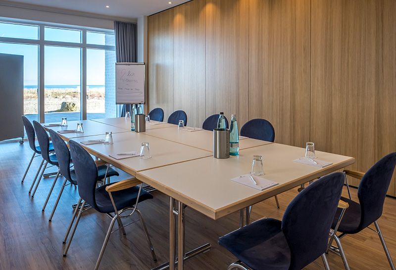 Tagen am Meer, Tagungsraum mit Bestuhlung in Block-Form, blaue Stühle, Tische eingedeckt mit Gläsern, Wasserflaschen, Schreibblöcke und Stift, an der Front Flipchart, Fensterfront mit Blick auf die Sonnenterrasse und Ostsee