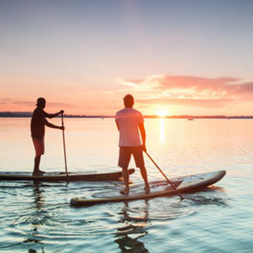 zwei Personen in kurzen Hosen beim Stand-up-Paddling, auf dem Meer bei Abendstimmung, im Hintergrund Sonnenuntergang
