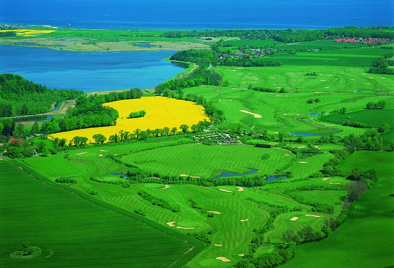 Golfplatz Hohwachter Bucht, Sicht von oben auf den kompletten Golfplatz, im Vordergrund die Golfflächen, links daneben blühendes gelbes Rapsfeld, im Hintergrund Ostsee Hohwachter Bucht