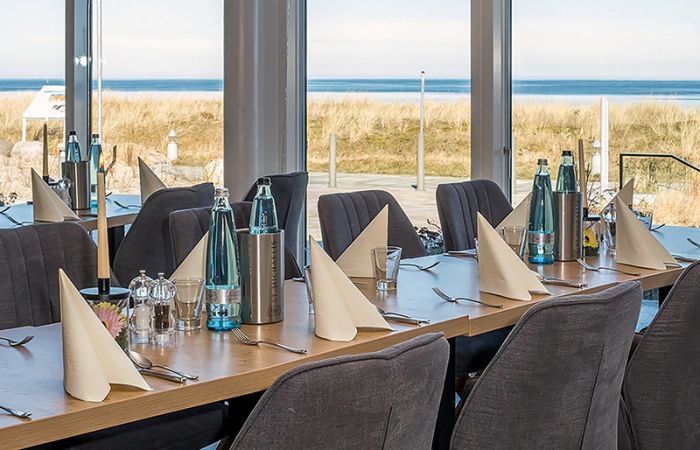 gedeckter Tisch im Restaurant mit grauen Sitzmöbeln, im Hintergrund Blick auf Sonnenterrasse und Meer