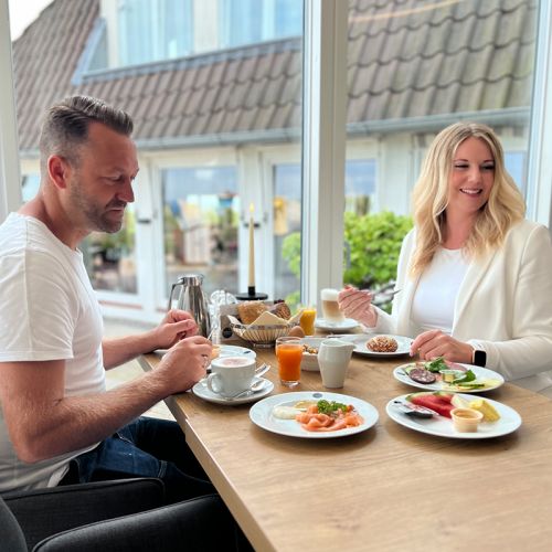 Restaurant Hotel Haus am Meer, Pärchen am gedeckten Tisch beim Frühstücken am Fenster, Frau schaut lächelnd in den Raum, Mann schaut auf den gedeckten Tisch