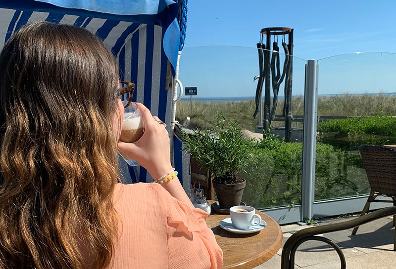 Restaurant Hotel Haus am Meer, Sonnenterrasse, Strandkorb und Rattanstuhl zum Sitzen, im Vordergrund Rücken einer Person, Person trinkt Kaffee, im Hintergrund Ostsee