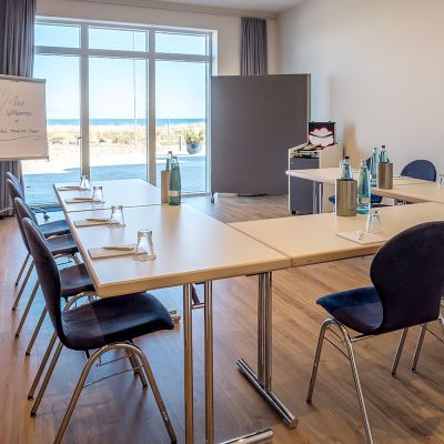 Tagen am Meer, Tagungsraum mit Bestuhlung in U-Form, Tische eingedeckt mit Gläsern, Wasserflaschen, Schreibblöcke und Stift, an der Front, Pinnwand auf Füßen, Plipchart, Rollwagen mit Tagungsutensilien, Fensterfront mit Blick auf Sonnenterrasse und Ostsee