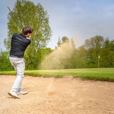 Golfplatz Hohwachter Bucht, Neudorfer Platz, im Vordergrund Golfer versucht Ball aus Sandfläche zu schlagen, aufgewirbelter Sand, vor dem Golfer das Grün mit Loch und Fähnchen, im Hintergrund Baumwand