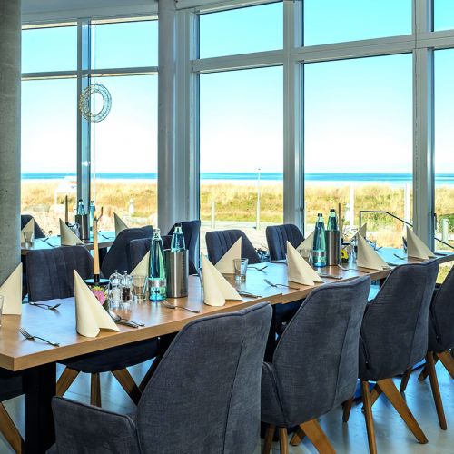Feierlichkeiten Haus am Meer, blaue Stühle an eingedeckter Tafel in der Lobby am Fenster, Besteck, Gläser, gefaltete Servietten, Wasserflaschen, Blick auf die Sonnenterrasse