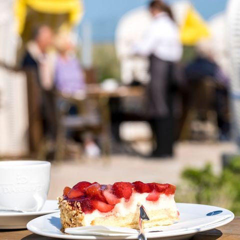 Restaurant Hotel Haus am Meer, im Vordergrund Kaffeetasse und Teller mit einem Stück Erdbeertorte, im Hintergrund verschwommen Strandkorb mit Paar und Bedienung