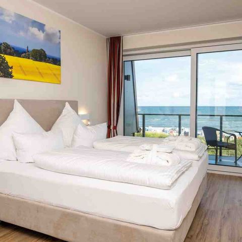 Hotel Haus am Meer, Appartement, Schlafzimmer, Boxspringbett, auf dem Bett liegen Bademantel und Badelatschen, über dem Bett Leinwand mit Motiv Rapsfeld, Balkon mit Blick auf Ostsee