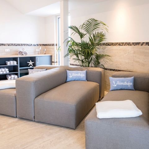 Wellness Hotel Haus am Meer, Wellnessbereich, graue Sofa mit Kissen und Decke, hinter Sofas Grünpflanze, im Hintergrund Sideboard mit Handtüchern