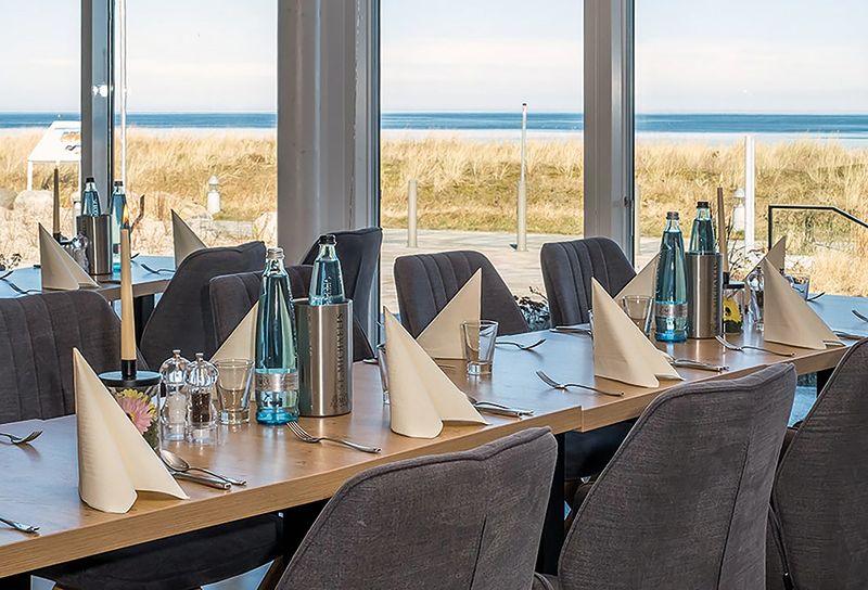 gedeckter Tisch im Restaurant mit grauen Sitzmöbeln, im Hintergrund Blick auf Sonnenterrasse und Meer