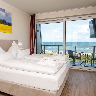 Appartements Hotel Haus am Meer, Schlafzimmer, Boxspringbett, auf dem Bett liegen Bademantel und Badelatschen, über dem Bett Leinwand mit Motiv Rapsfeld, Balkon mit Blick auf Ostsee