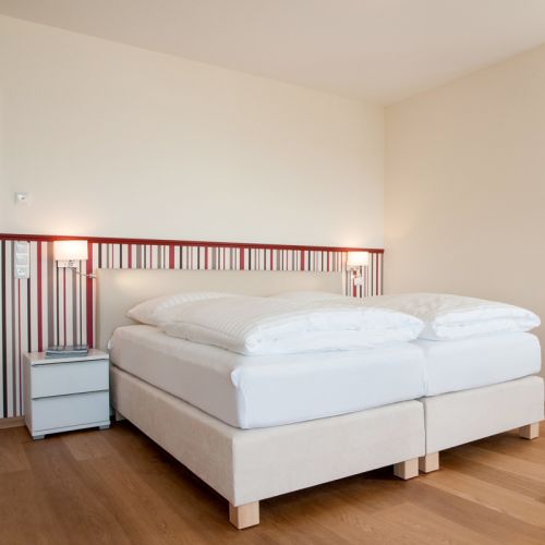 Schlafzimmer des Doppelzimmers. Neben dem Bett stehen Nachtische und kleine Lampen hängen an der Wand darüber