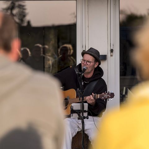 Aktivitäten Hohwachter Bucht, Chill at the Beach, Mann mit Brille und Hut sitzt auf Stuhl mit Gitarre, singt ins Mikrofon, im Vordergrund verschwommene Zuschauer