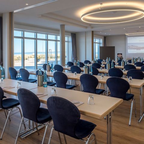 Tagen Hotel Haus am Meer, Tagungsraum, Bestuhlung Parlament, Tische eingedeckt mit Gläsern, Wasserflaschen, Schreibblöcken und Stift, an der Front Leinwand, auf der linken Seite Fensterfront mit Blick auf da Meer