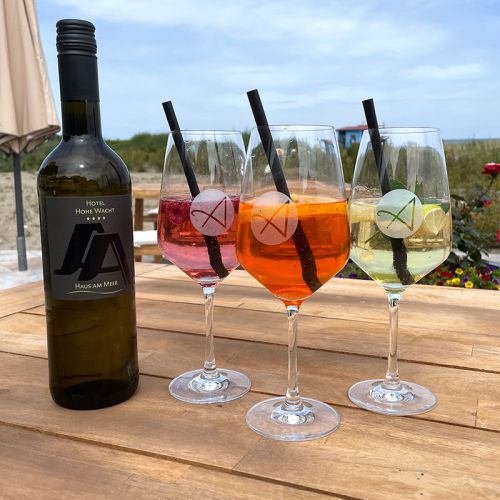 Restaurant Hotel Haus am Meer, Sonnenterrasse Holztisch, auf dem Tisch Weinflasche, drei Gläser mit Cocktails, im Hintergrund Sicht auf Düne und Wasser