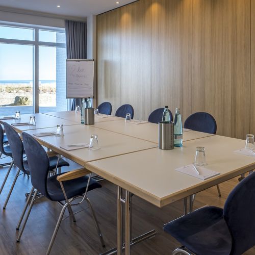 Tagen am Meer, Tagungsraum mit Bestuhlung in Block-Form, blaue Stühle, Tische eingedeckt mit Gläsern, Wasserflaschen, Schreibblöcke und Stift, an der Front Flipchart, Fensterfront mit Blick auf die Sonnenterrasse und Ostsee