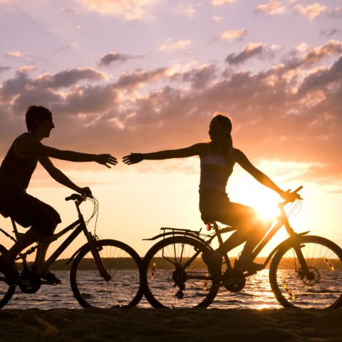 Aktivitäten Hohwachter Bucht, Pärchen auf Fahrrädern fahren am Strand lang beim Sonnenuntergang, Frau dreht sich um und reicht dem Mann die Hand