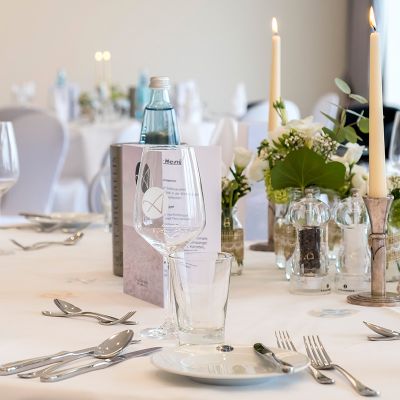 Feierlichkeiten Haus am Meer, festlich geschmückter Tisch, Teller, Besteck, Gläser mit Logo, gefaltete Servietten, Kerzen, Salz- und Pfefferstreuer, aufgeklappte Menükarte, Wasserflasche, Vase, Blumendeko