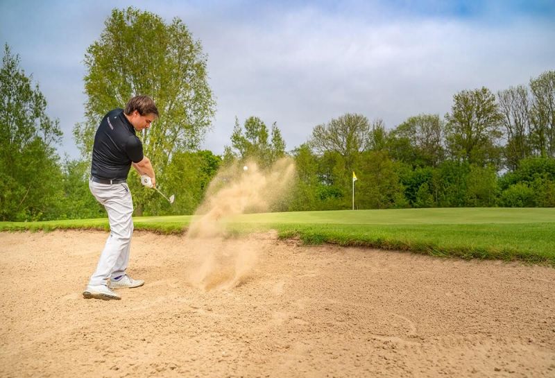Golfplatz Hohwachter Bucht, Neudorfer Platz, im Vordergrund Golfer versucht Ball aus Sandfläche zu schlagen, aufgewirbelter Sand, vor dem Golfer das Grün mit Loch und Fähnchen, im Hintergrund Baumwand