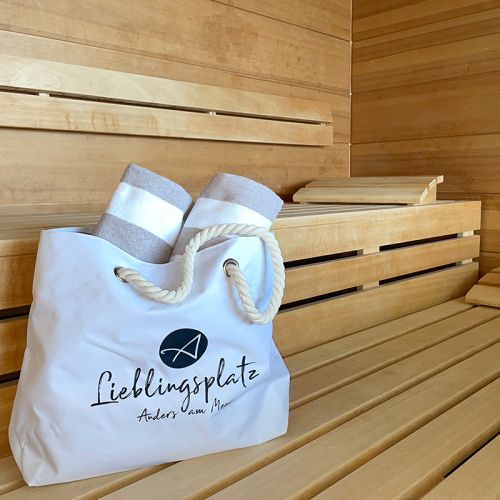 Tageswellness „Genuss am Meer“ Hohe Wacht, Sauna, weiße Wellnesstasche mit Kordelhänkel, auf der Tasche Logo Aners am Meer und Aufdruck Lieblingsplatz, aus der Tasche ragen zwei weiß-beige gestreifte eingerollte Handtücher, Tache steht auf der Holzbank