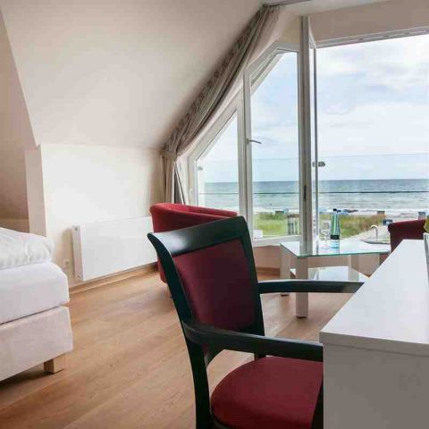Zimmer Hotel Haus am Meer, Hotelzimmer mit Boxspringbett, Schreibtisch mit Stuhl und Sitzecke mit Sessel und Tisch, große Fensterfront, geöffnete Terrassentür mit Blick aufs Meer