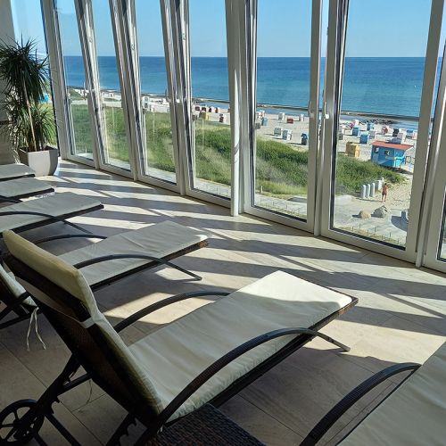 Hotel Haus am Meer, Wellnessbereich, Wellnessliegen in einer Reihe mit Blick durch die Fensterfront auf die Ostsee, links in der Ecke große grüne Topfpflanze, Blick durch die Fensterfront auf den Strand und die Ostsee, Strandkörbe am Strand